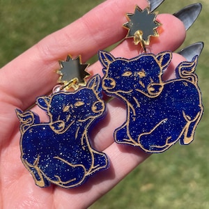 Könnte beinhalten: Ein Paar blaue Glitzer-Hirschohrringe mit goldenen Stern-Akzenten. Die Hirsche blicken nach vorne, ihre Köpfe sind leicht zur Seite geneigt. Die Ohrringe sind eine skurrile und lustige Ergänzung zu jedem Outfit.