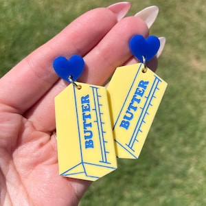 Könnte beinhalten: Ein Paar gelbe und blaue Ohrringe in Form von Butterstangen. Jeder Ohrring hat oben einen blauen Herzanhänger und das Wort "BUTTER" ist auf der Vorderseite aufgedruckt.