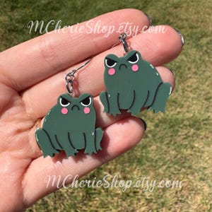 May include: A pair of green frog earrings with a grumpy expression. The frogs have white eyes, black eyebrows, and pink cheeks. The earrings are suspended from silver-colored hooks. The text "MCherieShop etsy.com" is visible.