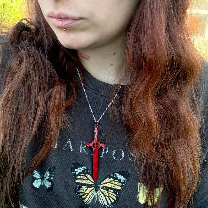 May include: A silver chain necklace with a red sword pendant. The sword has a cross-shaped guard and a pointed tip.