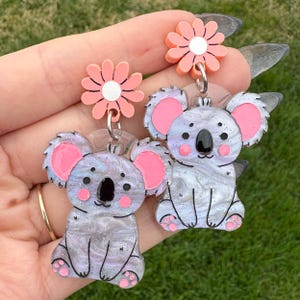 May include: A pair of earrings featuring a pink flower charm and a koala charm with a marbled grey and white finish. The koala charm has pink ears and a black nose.