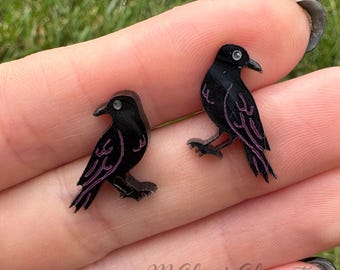 May include: A pair of black crow-shaped earrings with purple etched details. Each crow has a small, clear gem for an eye. The earrings are displayed against a blurred green background.