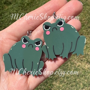May include: A pair of green frog-shaped earrings with black angry eyebrows and pink cheeks. The earrings have a silver-colored hook and the text "MCherieShop.etsy.com" is printed on the bottom of the earrings.