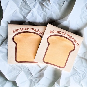 May include: Two square cards with a light brown toast illustration and the text "Breader Than You" in brown. The cards are on a white, crumpled paper background. The toast illustration has a brown outline.