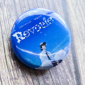 Pode incluir: Um botão redondo azul com o texto "Baekhyun World Tour" e "Revesie" em escrita branca. O botão apresenta uma imagem de uma pessoa com camisa branca e gravata contra um fundo de céu.