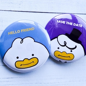 May include: Two round buttons with cartoon duck faces. One button is light blue with the text "HELLO FRIEND" in gold. The other button is purple with a duck wearing a top hat and the text "SAVE THE DATE" in white.