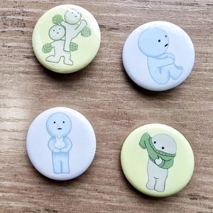 May include: Four round button badges featuring different illustrations of a simple white character with a blue face. The character is shown in various poses and situations, including holding a smaller version of itself, wearing a green jumper, and being held up by another character. The badges are on a light brown wooden surface.