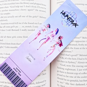 Puede incluir: Un marcapáginas lila y blanco con un diseño de entrada de concierto. El marcapáginas presenta tres figuras estilizadas, el texto "HUNTRAX WORLD TOUR" y las palabras "GONNA BE Golden". El logotipo de Huntrax está en la parte superior.