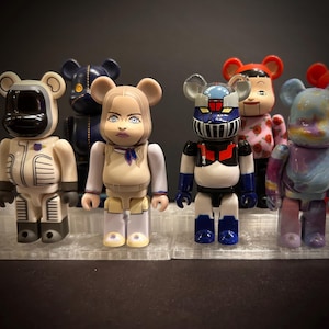 May include: A collection of colorful Bearbrick figures, each with unique designs. One figure is dressed as an astronaut, another as a robot, and others feature various patterns and characters. The figures are displayed on a clear, textured surface.