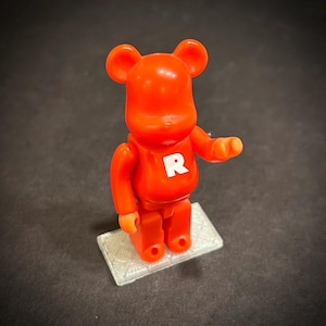May include: A red Bearbrick figure with a white "R" on its chest, standing on a small, rectangular platform. The figure has a bear-like head and articulated limbs. The background is a dark gray.