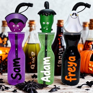 May include: Three plastic tumblers with lids and straws, each topped with a Halloween character: a purple vampire, a green Frankenstein monster, and a white ghost. The purple cup says Sam, the green cup says Adam, and the black cup says Freya.