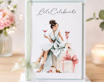 May include: A greeting card featuring an illustration of a person in a light blue dress sitting on gift boxes, holding a cocktail glass. The text "Let's Celebrate" is printed at the top. A sheer light green ribbon with a bow is attached to the side.