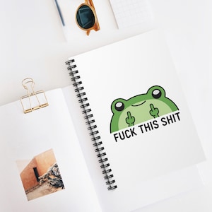 Peut inclure: Cahier blanc à spirales avec une grenouille verte de dessin animé faisant un doigt d'honneur et le texte "FUCK THIS SHIT". Le cahier est ouvert, avec une pince à reliure dorée et des lunettes de soleil visibles.