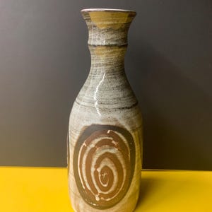 Peut inclure: Un vase en céramique avec un col étroit et un bord évasé, présentant un vernis gris et beige moucheté. Le corps du vase est orné d'un motif en spirale marron proéminent centré sur le devant, avec des accents jaunâtres près du sommet.