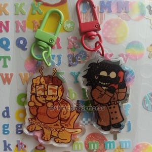 May include: Two acrylic keychains with cartoon illustrations. One features a dog with glasses and a dessert, the other a character in a trench coat. Both have colorful carabiner clips. The artist's name, @sketchingdemonss, is visible.