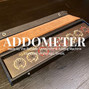 Peut inclure: Un étui en cuir noir et marron vintage contenant une machine à calculer en métal. La machine possède un cadran rouge et blanc et le texte "ADDOMETER Made by the Reliable Typewriter & Adding Machine Company of Chicago, Illinois." est imprimé sur l'étui.