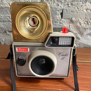 Puede incluir: Una cámara vintage Ansco Cadet II con un accesorio de flash dorado. La cámara es plateada y negra con una correa negra. La cámara tiene una lente, un visor y una perilla de avance de película. La cámara está etiquetada como "Ansco Cadet II" y "Use 127 Film".