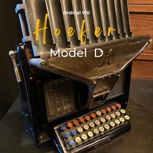 May include: An antique black and bronze Hoeffer Model D machine with a numerical keypad. The machine has a hinged top and a series of slots. The text "Ordinal VIII" and "Hoeffer" are in yellow script, and "Model D" is in white.