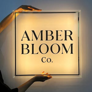 May include: A square, illuminated sign with the text "AMBER BLOOM Co." in black, sans-serif font. The sign is held by two hands, with a warm, yellow glow emanating from within. The sign has a clear border.