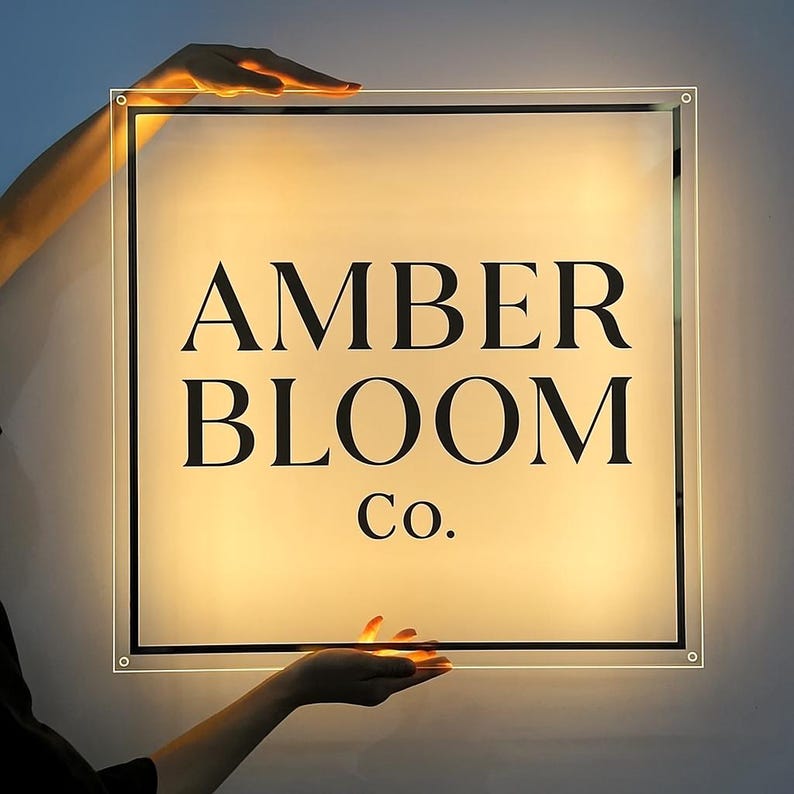 May include: A square, illuminated sign with the text "AMBER BLOOM Co." in black, sans-serif font. The sign is held by two hands, with a warm, yellow glow emanating from within. The sign has a clear border.