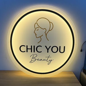 May include: A round, illuminated sign with the text "CHIC YOU Beauty" in black and gold. The sign features a gold outline of a woman's face and a black border. The background is a warm, glowing yellow.