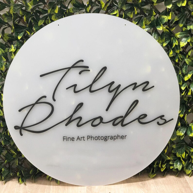 May include: A white, circular sign with the name "Tilyn Rhodes" in elegant black script. Below the name, the text reads "Fine Art Photographer" in a smaller, sans-serif font. The sign has two small holes at the top.