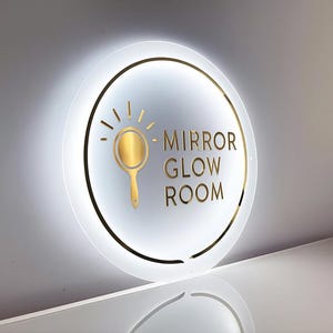 May include: Round, illuminated sign with the words "MIRROR GLOW ROOM" in gold lettering. A gold-colored mirror icon is centered above the text. The sign has a gold border and is set against a white background.