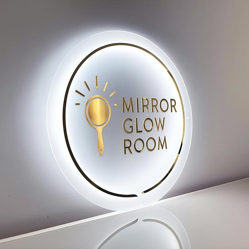 May include: Round, illuminated sign with the words "MIRROR GLOW ROOM" in gold lettering. A gold-colored mirror icon is centered above the text. The sign has a gold border and is set against a white background.