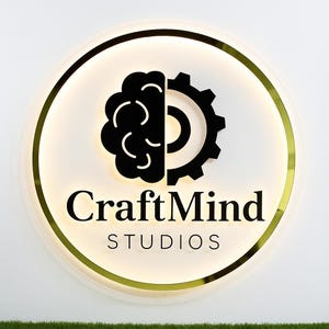 May include: A circular wall sign with a gold metallic rim and backlighting. The center features a black logo combining a brain and a gear, with the text CraftMind STUDIOS printed in black below the icon on a white background.