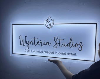 May include: A rectangular sign with the text "Wynterin Studios" in a cursive font, with the tagline "Cool elegance shaped in quiet detail." The sign has a floral design above the text and is illuminated with a soft, white light.