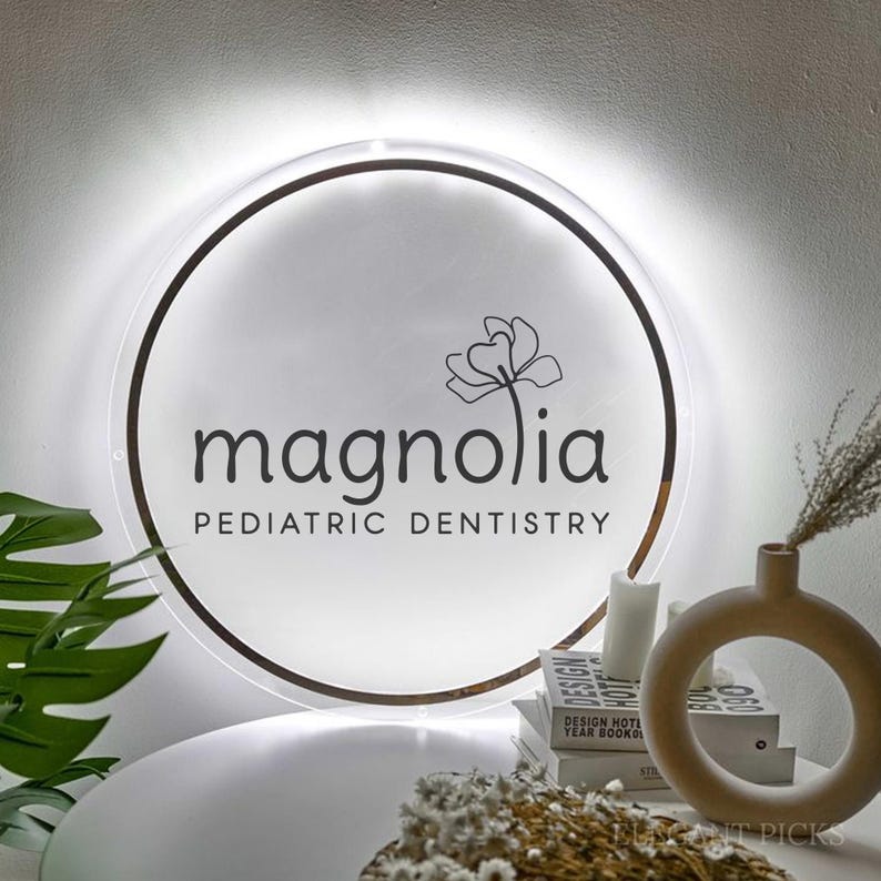 May include: A round, illuminated sign with the text "magnolia PEDIATRIC DENTISTRY" in black. A magnolia flower graphic is above the text. The sign is surrounded by a brown ring and white light. The sign is on a white surface.