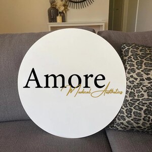 May include: A white, round sign with the word "Amore" in black script, and "Medical Aesthetics" in gold script. The sign is resting on a gray sofa, with a leopard print pillow visible.