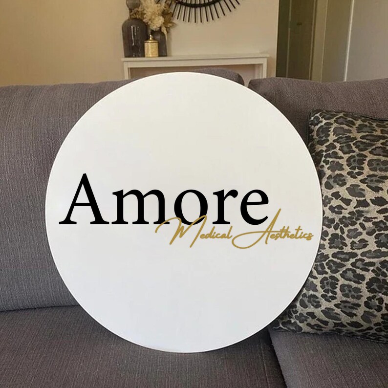 May include: A white, round sign with the word "Amore" in black script, and "Medical Aesthetics" in gold script. The sign is resting on a gray sofa, with a leopard print pillow visible.