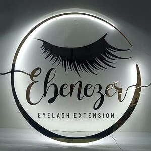 May include: A circular illuminated sign with the name "Ebenezer" in elegant script, featuring a stylized eyelash design. The words "EYELASH EXTENSION" are below the name, all against a white background.