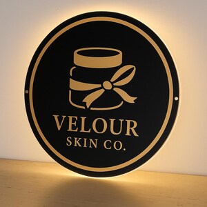 Op de afbeelding: Een ronde, zwarte plaat met een gouden rand en een gouden illustratie van een pot met een strik. De tekst "VELOUR SKIN CO." staat in gouden letters eronder. Het bord wordt verlicht door een warm, zacht licht.