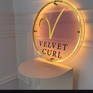 May include: A round sign with the text "VELVET CURL" in black, and a gold-colored checkmark symbol. The sign is illuminated with a warm light and mounted on a white shelf. The sign has a gold-colored border.