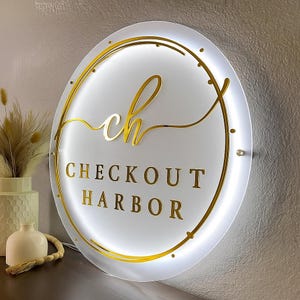 Op de afbeelding: Een rond, verlicht bord met de tekst "CHECKOUT HARBOR" in gouden letters. Het bord heeft een gouden rand en de gestileerde letters "ch" in het midden. Het bord is wit en heeft een zachte gloed.