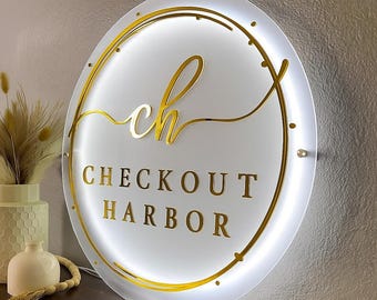 May include: A circular white acrylic sign featuring gold lettering that reads "ch" in script and "CHECKOUT HARBOR" in block font. The sign is framed by a gold circular border and illuminated by a bright white LED light ring.