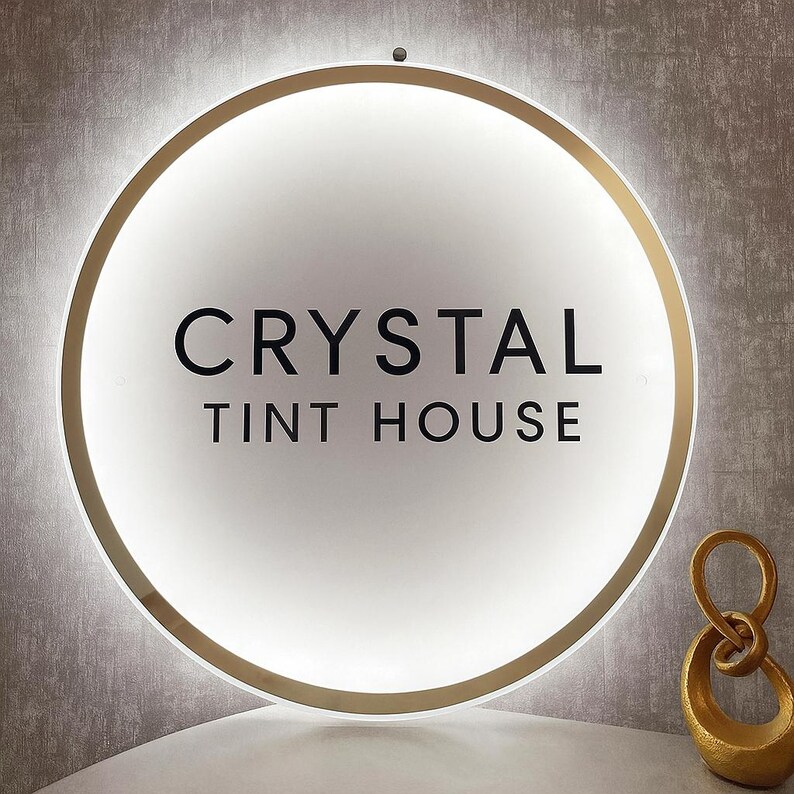 May include: A round, illuminated sign with the text "CRYSTAL TINT HOUSE" in black. The sign has a gold-colored border and a white, glowing center. A gold-colored decorative sculpture is to the right of the sign.