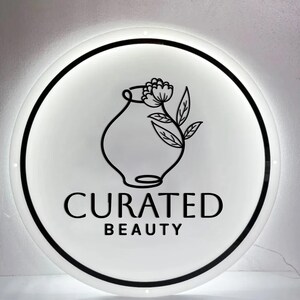 Puede incluir: Un letrero redondo e iluminado con una cara blanca y un borde negro. El letrero presenta un dibujo lineal negro de un jarr&oacute;n con una flor y hojas. Las palabras "CURATED BEAUTY" est&aacute;n impresas en negro debajo.