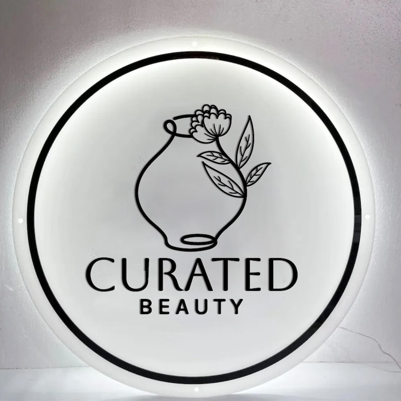 Puede incluir: Un letrero redondo e iluminado con una cara blanca y un borde negro. El letrero presenta un dibujo lineal negro de un jarr&oacute;n con una flor y hojas. Las palabras "CURATED BEAUTY" est&aacute;n impresas en negro debajo.