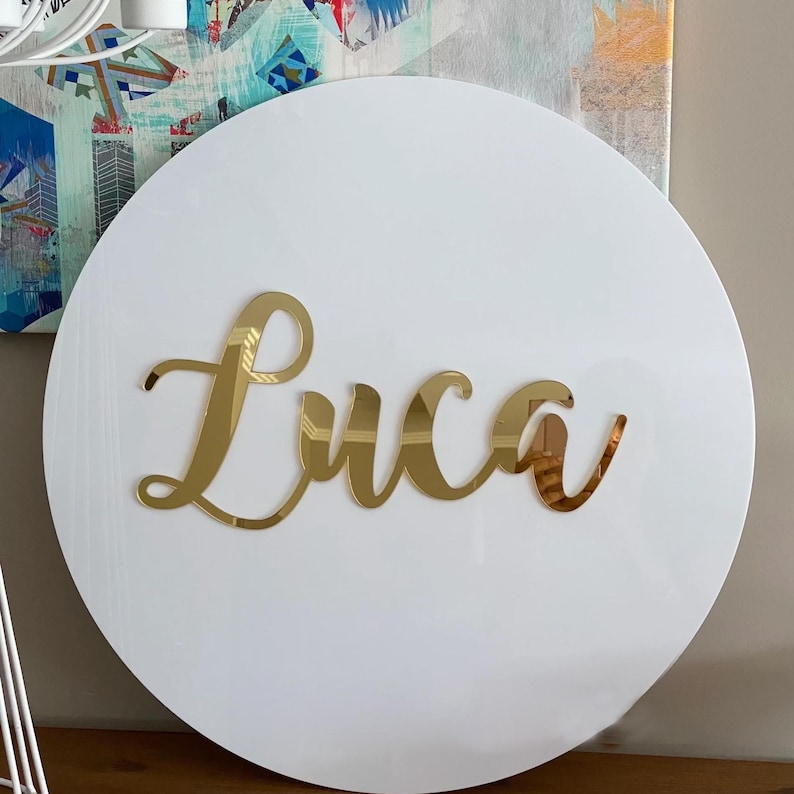 May include: A white, circular wooden sign with the name "Luca" in gold, cursive lettering. The sign is a decorative piece, likely for a nursery or child's room. The background includes a colorful abstract painting.