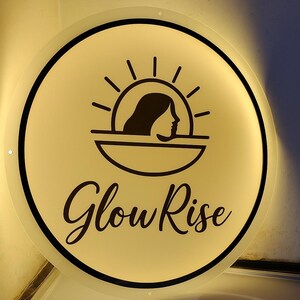 Puede incluir: Un letrero redondo e iluminado con una cara color crema y un borde negro. El letrero presenta un gr&aacute;fico negro del perfil de una mujer debajo de un dise&ntilde;o de sol, con las palabras "Glow Rise" en una escritura elegante.