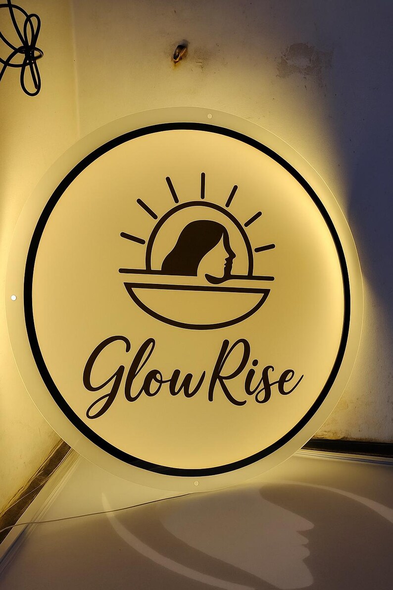 Puede incluir: Un letrero redondo e iluminado con una cara color crema y un borde negro. El letrero presenta un gr&aacute;fico negro del perfil de una mujer debajo de un dise&ntilde;o de sol, con las palabras "Glow Rise" en una escritura elegante.