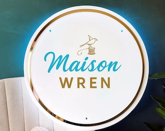 May include: A round, white sign with a gold rim and a teal blue glow. The sign features the words "Maison WREN" in teal and gold lettering, with a gold bird and spool graphic above the word "WREN".
