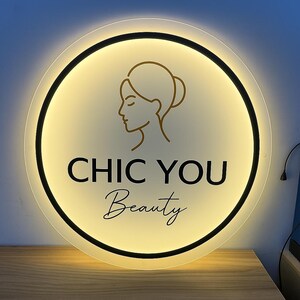 May include: A round, illuminated sign with the text "CHIC YOU Beauty" in black and gold. The sign features a gold outline of a woman's face and a black border. The background is a soft, warm yellow.
