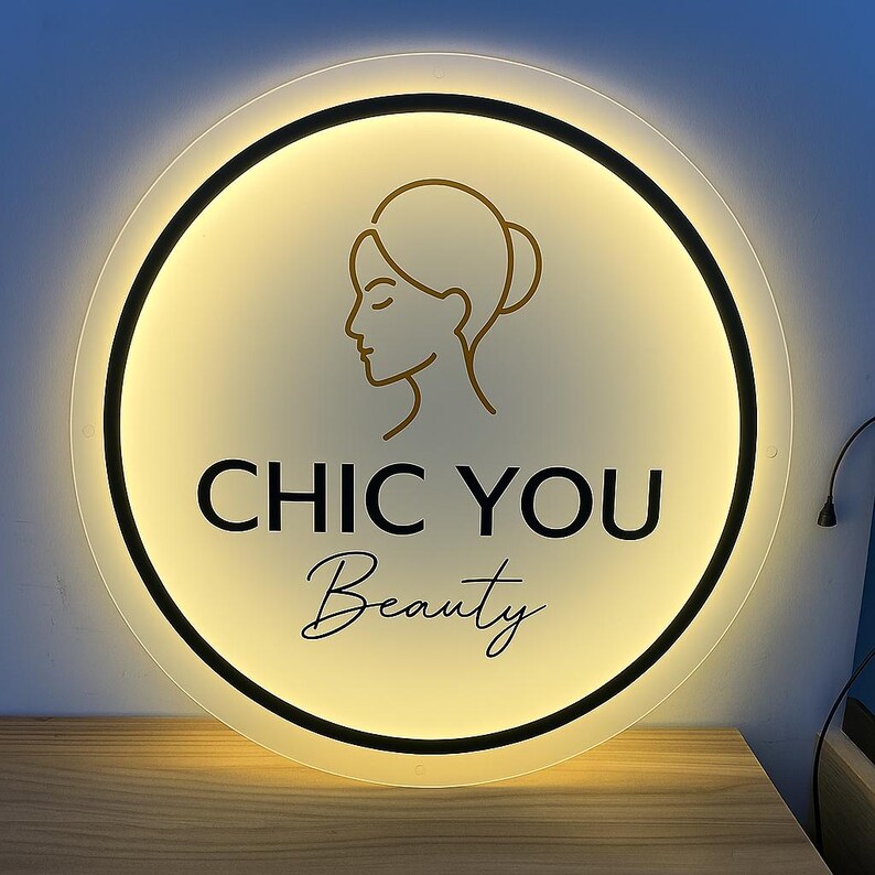 May include: A round, illuminated sign with the text "CHIC YOU Beauty" in black and gold. The sign features a gold outline of a woman's face and a black border. The background is a soft, warm yellow.