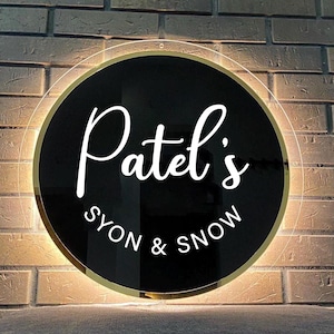 May include: A circular black acrylic sign with a gold-colored border and backlighting. The sign features the white text "Patel's" in a large script font, with the text "SYON & SNOW" in a smaller, clean sans-serif font below it.
