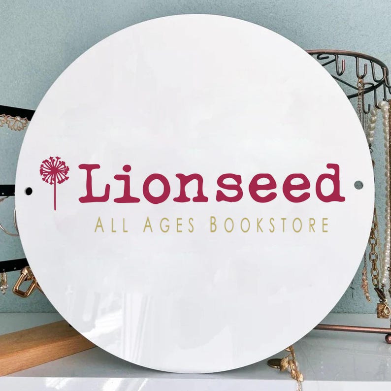 May include: A white, round sign with the text "Lionseed ALL AGES BOOKSTORE" in burgundy and gold lettering. A dandelion graphic is on the left side. The sign is displayed in front of a jewelry display.