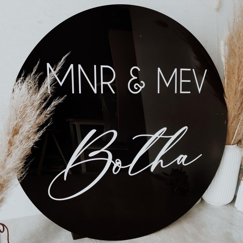 May include: A round, black acrylic sign with white lettering. The text reads "MNR & MEV" above "Botha" in a cursive font. The sign is a decorative item, possibly for a wedding or event.